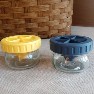 May include: Two glass jars with lids. The jar on the left has a yellow lid with a segmented design. The jar on the right has a blue lid with a segmented design.