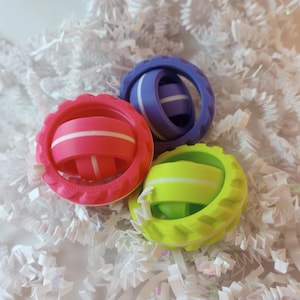 May include: Three fidget toys in different colors: pink, purple, and green. Each toy has a white stripe around the center and is made of plastic.