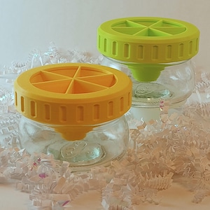 May include: Two clear glass jars with colorful lids. One jar has a yellow lid, and the other has a green lid. The lids have a circular design with vertical slots around the edge and a cross-shaped pattern on top. The jars are sitting on white paper.