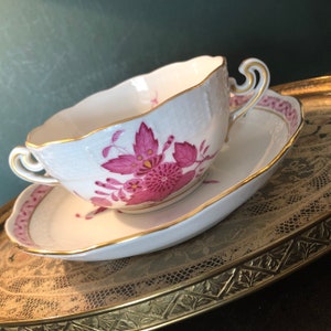 May include: A white porcelain teacup and saucer with a pink floral design. The cup has a scalloped edge and a gold rim. The saucer has a gold rim and a pink floral design around the edge.
