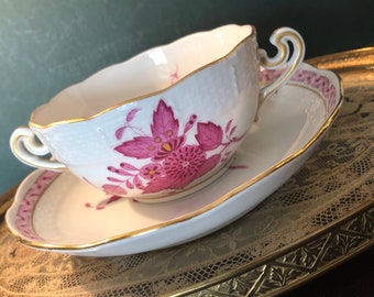 May include: A white porcelain teacup and saucer with a pink floral design. The cup has a scalloped edge and a gold rim. The saucer has a gold rim and a pink floral design around the edge.