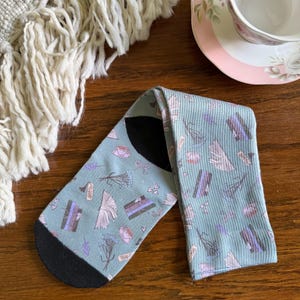 May include: A pair of light blue socks with a black heel and toe. The socks feature a pattern of books, flowers, teacups, and bows in purple, pink, and white. The socks are folded on a wooden surface, with a white fringed blanket and a teacup in the background.