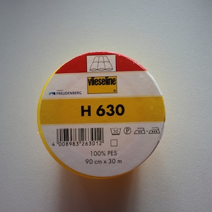Può includere: Un rotolo di interfodera termoadesiva Vlieseline H 630 giallo e rosso. L'etichetta include il nome del marchio, il numero del prodotto, le istruzioni di cura e il testo "100% PES 90 cm x 30 m".