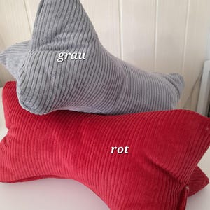 以下が含まれることがあります： グレーと赤の2つの首枕。コーデュロイ生地で作られています。枕は三日月型で、中央が盛り上がっています。グレーの枕には「grau」と書いてあり、赤い枕には「rot」と書いてあります。