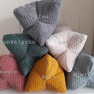 Könnte beinhalten: Sechs Plüschkissen in Sternform in verschiedenen Farben, darunter Grau, Dunkelgrün, Mauve, Gelb, Mint und Beige. Die Kissen sind mit den Farben "grau", "dunkelgrün", "mauve", "mais", "mint" und "natur" beschriftet.