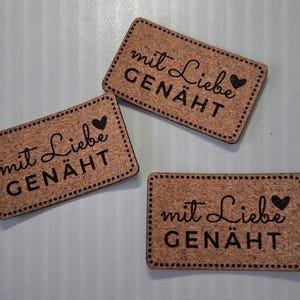 Könnte beinhalten: Drei rechteckige Korketiketten mit abgerundeten Ecken, jedes mit einem gestickten Randdesign und dem schwarz lasergravierten Text "mit Liebe GENÄHT" sowie einem kleinen Herzsymbol, angeordnet in einem Streumuster auf einer hellen Fläche.