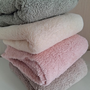 Op de afbeelding: Een stapel van vier opgevouwen pluche dekens in verschillende neutrale kleuren. De dekens zijn grijs, gebroken wit, roze en taupe. Het zachte, getextureerde materiaal is zichtbaar in de close-up. De dekens zijn netjes op elkaar gestapeld.