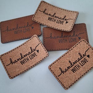 Puede incluir: Cinco etiquetas rectangulares con esquinas redondeadas, que presentan una mezcla de acabados en marrón liso y corcho texturizado. Cada etiqueta está marcada con el texto negro "handmade WITH LOVE" y un pequeño corazón, con borde cosido.