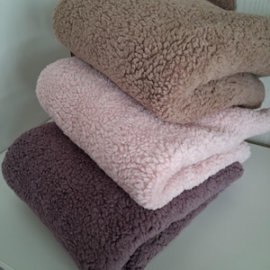 Op de afbeelding: Een stapel van vier pluche dekens in verschillende tinten bruin, roze en paars. De dekens hebben een zachte, getextureerde uitstraling, wat een gezellig en comfortabel gevoel suggereert. De dekens zijn netjes opgevouwen en tonen hun zachte textuur.