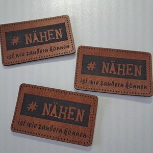 Peut inclure: Trois étiquettes rectangulaires marron avec des bordures marron foncé et des détails de couture. Chaque étiquette présente le texte #NÄHEN en police grasse, suivi de la phrase "ist wie zaubern können" dans une police cursive plus petite.