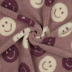 Puede incluir: Primer plano de una tela suave y texturizada en tonos malva y crema. La tela está estampada con caras sonrientes repetidas en crema y morado oscuro. El material parece afelpado y acogedor, ideal para mantas o ropa.