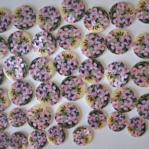 May include: A collection of round buttons with a floral design. The buttons feature a cream-colored background with a painted image of pink and purple flowers. The buttons vary in size and are arranged on a white surface.