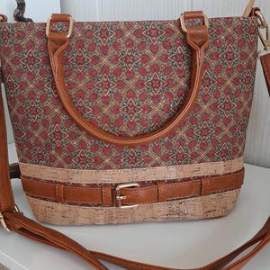 May include: A brown cork and fabric handbag with a geometric pattern and a brown leather strap. The bag has a buckle closure and a top handle.