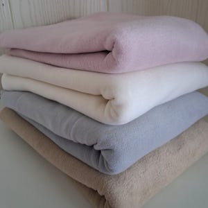 Könnte beinhalten: Ein Stapel von fünf gefalteten Fleece-Decken in Pastellfarben. Die Decken sind in Rosa, Weiß, Hellblau und Beige gehalten. Die Decken sind ordentlich gefaltet und übereinander gestapelt.