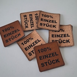 Puede incluir: Un conjunto de etiquetas cuadradas de color marrón y beige con textura, que presentan el texto impreso en negro 100% EINZEL STÜCK. Cada etiqueta tiene un diseño de borde cosido y están dispuestas de forma dispersa y superpuesta.