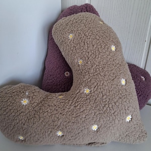 May include: Two heart-shaped decorative cushions. The front cushion is light brown with small white and yellow daisy embroidery. The back cushion is a solid purple. Both cushions have a textured, fuzzy appearance.