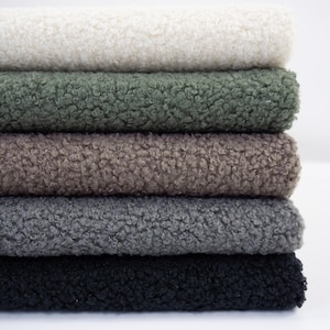 Könnte beinhalten: Ein Stapel aus fünf gefalteten Stücken Sherpa-Fleece-Stoff. Die Farben von oben nach unten sind Creme, Olivgrün, Taupe, Anthrazitgrau und Schwarz. Jedes Stück weist eine dicke, lockige und strukturierte Floroberfläche mit einheitlichem Aussehen auf.
