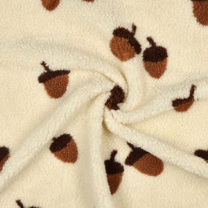 Puede incluir: Tela color crema con un patrón repetido de bellotas marrones. Las bellotas tienen un aspecto texturizado y la tela tiene una textura suave y esponjosa. Este material acogedor es adecuado para mantas, mantas o otros artículos de decoración del hogar.