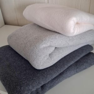 Op de afbeelding: Een stapel van vier gevouwen fleece dekens in verschillende tinten grijs en wit. De bovenste deken is een licht off-white, gevolgd door lichtgrijs, middengrijs en een donker antracietgrijs aan de onderkant, allemaal netjes opgevouwen.