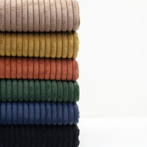 May include: A stack of colorful corduroy fabric in shades of tan, gold, rust, green, navy, and black. The fabric has a ribbed texture and is neatly folded, showcasing the variety of colors available for sewing or crafting projects.