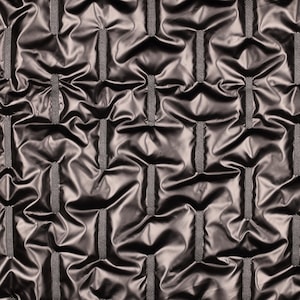 Puede incluir: Una tela gris metálico oscuro con un patrón repetitivo de tiras verticales tejidas y texturizadas que crean pliegues profundos y fruncidos, dando una apariencia acolchada a toda la superficie del material con un acabado altamente reflectante.