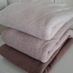 Könnte beinhalten: Ein Stapel aus vier gefalteten Decken in verschiedenen neutralen Farbtönen. Die Decken reichen von hellem Beige bis zu einem tiefen Braun, wodurch ein Farbverlauf entsteht. Der Stoff wirkt weich und flauschig und suggeriert Wärme und Komfort.