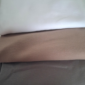 Peut inclure: Trois couches de tissu plié de couleurs unies : blanc, beige et gris. Le tissu semble être une matière douce, peut-être du coton ou un mélange similaire. Les plis créent un effet de superposition, mettant en valeur les différentes teintes.