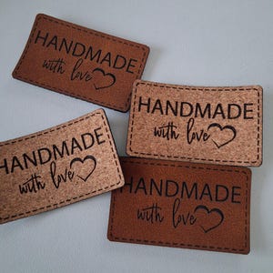 Puede incluir: Cuatro etiquetas rectangulares con esquinas redondeadas, con el texto HANDMADE with love y un símbolo de corazón. Dos etiquetas son de material marrón liso y dos son de material de corcho marrón claro texturizado con costuras.