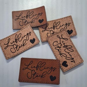 Peut inclure: Cinq étiquettes rectangulaires marron aux coins arrondis, chacune portant le texte noir "Lieblings Stück" en écriture cursive et une petite icône de cœur noir. Les étiquettes ont une surface texturée et des coutures visibles sur les bords.