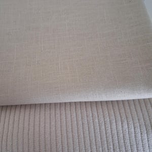 Può includere: Due campioni di tessuto in tonalità neutre. Il campione superiore ha una trama tessuta, mentre il campione inferiore presenta una trama a coste con righe verticali. Entrambi i tessuti sono di colore beige chiaro, adatti a vari progetti tessili.
