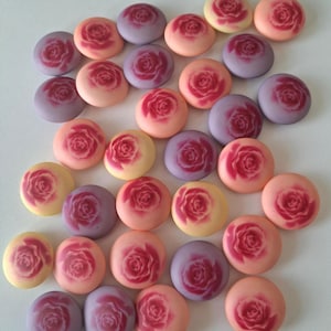 May include: A collection of round, pastel-colored candies, each adorned with a detailed rose design. The candies feature shades of pink, purple, and yellow, creating a visually appealing assortment. The rose details are in a darker shade of pink.