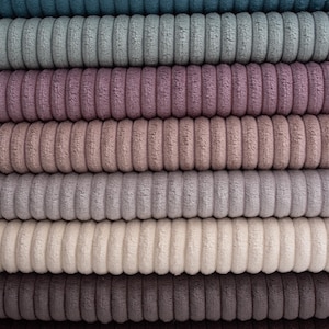 Op de afbeelding: Een stapel corduroy stof in verschillende kleuren, waaronder teal, grijs, lavendel, beige en bruin. De stof heeft een geribbelde textuur, waarbij elke kleurellaag netjes op elkaar is gestapeld. De corduroy ziet er zacht en pluche uit.