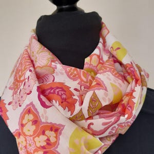 May include: A lightweight infinity scarf featuring a white base with a vibrant floral and paisley pattern in shades of bright pink, coral, orange, and lime green. The fabric is draped in soft folds, showcasing the repeating botanical print throughout.