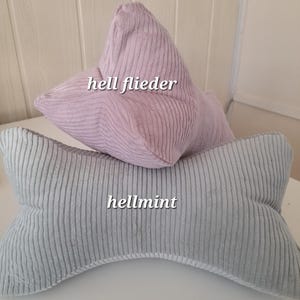 May include: Two corduroy pillows, one light pink and one light gray. The pink pillow is shaped like a star and has the text "hell flieder" on it. The gray pillow is shaped like a bone and has the text "hellmint" on it.