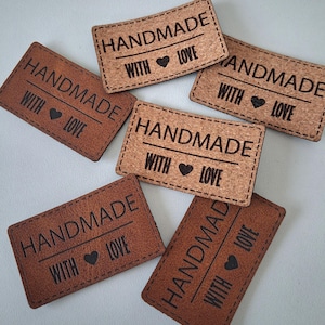 May include: A collection of rectangular labels in varying shades of brown, including cork and leather-like textures. Each label features the black engraved text HANDMADE WITH LOVE and a small heart symbol, with visible stitching along the edges.