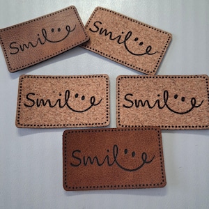 May include: Five rectangular labels with rounded corners and stitched edges. Three labels are made of light brown cork material, and two are made of smooth brown leather. Each label features the word "smile" laser-engraved in black cursive text.
