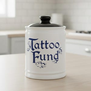 May include: A white ceramic jar with a black lid, featuring the words "Tattoo Fund" in blue script. The jar is cylindrical and sits on a light-colored wooden surface. The background shows a kitchen setting.