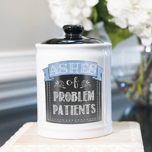 May include: White ceramic jar with a black lid and a blue and black chalkboard design that says "Ashes of Problem Patients".