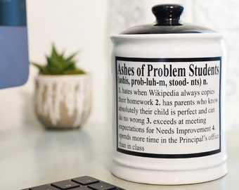 May include: White ceramic jar with a black lid and the text "Ashes of Problem Students (ashis, prob-luh-m, stood-nts) n. 1. hates when Wikipedia always copies their homework 2. has parents who know absolutely their child is perfect and can do no wrong 3. exceeds at meeting expectations for Needs Improvement 4. spends more time in the Principal's office than in class" printed on the front.