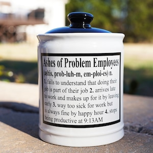 May include: White ceramic jar with a black lid. The jar has a black label with the text "Ashes of Problem Employees (ashis, prob-luh-m, em-ploi-es) n. 1. fails to understand that doing their job is part of their job 2. arrives late to work and makes up for it by leaving early 3. way too sick for work but is always fine by happy hour 4. stops being productive at 9:13AM"
