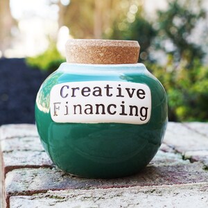 May include: A green ceramic jar with a cork lid. The jar has a white label with the text "Creative Financing" on it.