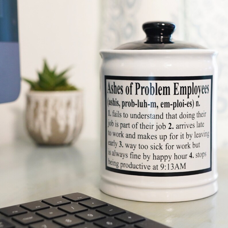 May include: White ceramic jar with a black lid. The jar has a black and white label that reads "Ashes of Problem Employees (ashis, prob-luh-m, em-ploi-es) n. 1. fails to understand that doing their job is part of their job 2. arrives late to work and makes up for it by leaving early 3. way too sick for work but is always fine by happy hour 4. stops being productive at 9:13AM"