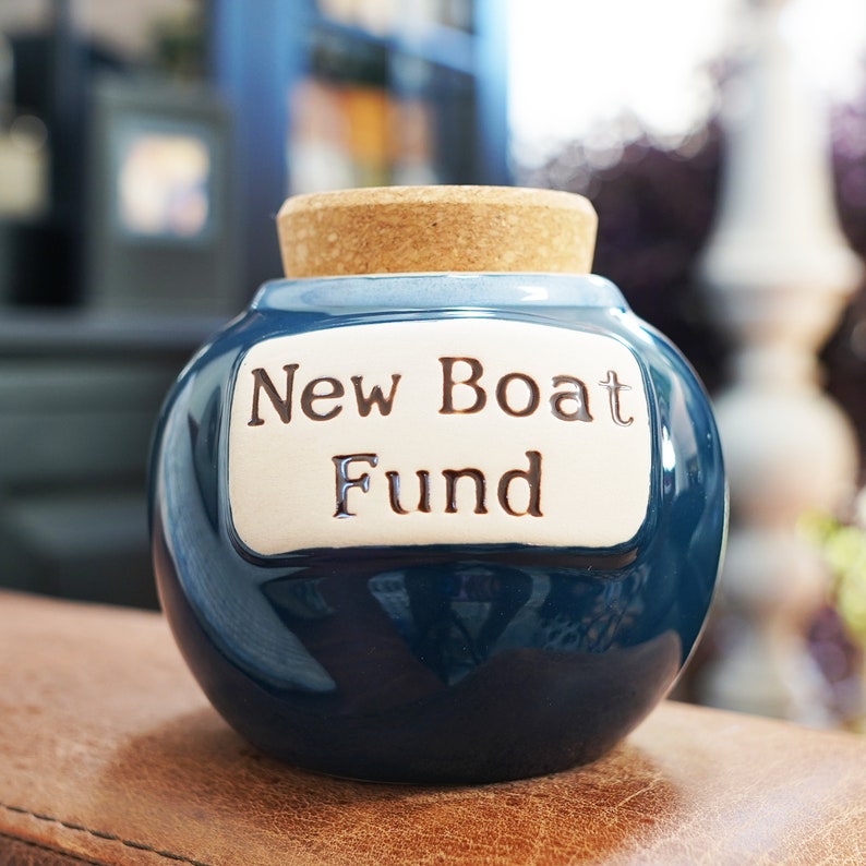 May include: A blue ceramic money box with a cork top and a white label that reads "New Boat Fund".