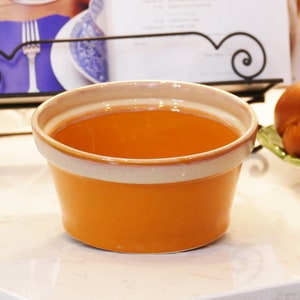 May include: A large, orange ceramic bowl with a white rim. The bowl is empty and sitting on a white countertop.
