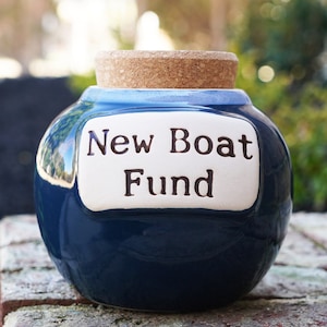 May include: A blue ceramic money box with a cork lid. The box has a white label that reads "New Boat Fund".
