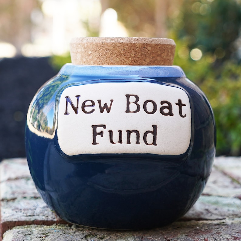 May include: A blue ceramic money box with a cork lid. The box has a white label that reads "New Boat Fund".