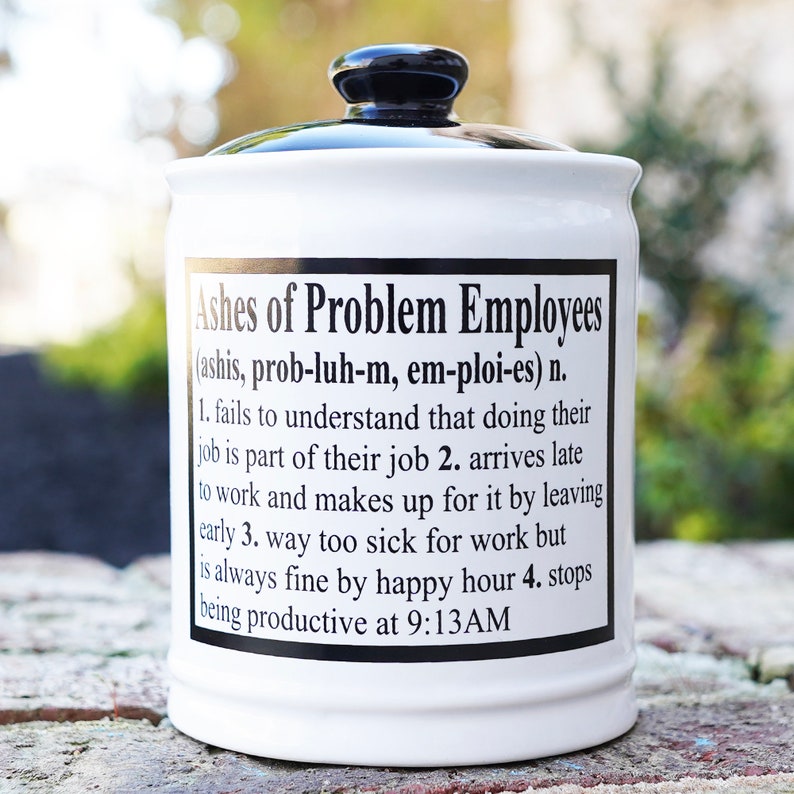 May include: White ceramic jar with a black lid. The jar has a black and white label that reads "Ashes of Problem Employees (ashis, prob-luh-m, em-ploi-es) n. 1. fails to understand that doing their job is part of their job 2. arrives late to work and makes up for it by leaving early 3. way too sick for work but is always fine by happy hour 4. stops being productive at 9:13AM"