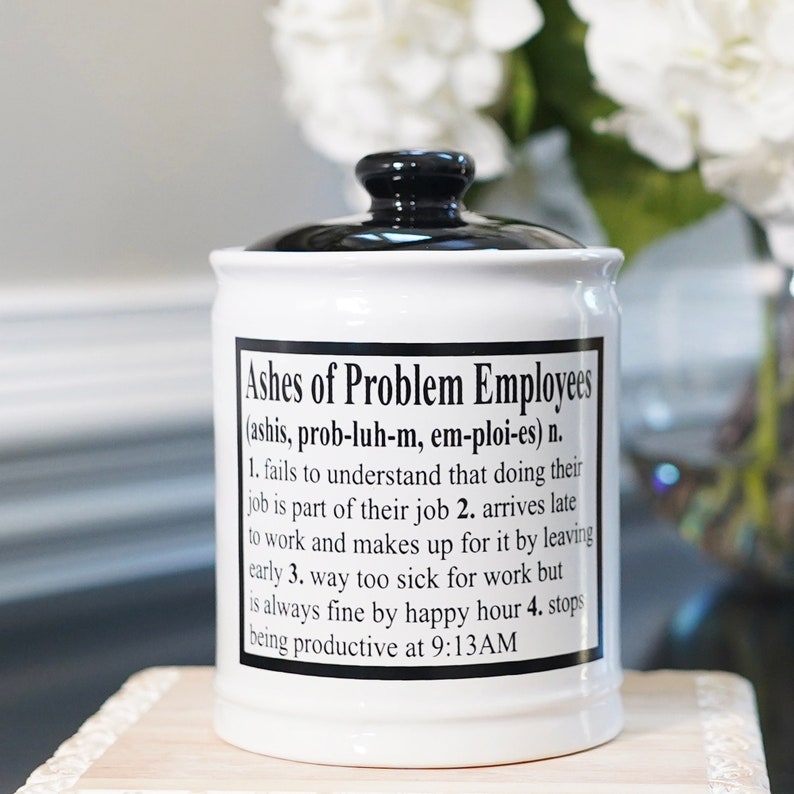 May include: White ceramic jar with a black lid. The jar has a black and white label that reads "Ashes of Problem Employees (ashis, prob-luh-m, em-ploi-es) n. 1. fails to understand that doing their job is part of their job 2. arrives late to work and makes up for it by leaving early 3. way too sick for work but is always fine by happy hour 4. stops being productive at 9:13AM"