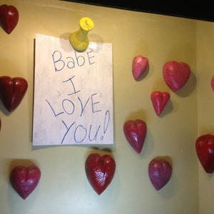 May include: A white card with the text "Babe I Love You!" written in blue ink is pinned to a wall with a yellow pushpin. The wall is decorated with several pink heart-shaped decorations.