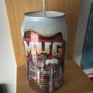 May include: A candle in the shape of a Mug Root Beer can. The can is brown and silver with the Mug Root Beer logo and text. The candle has a white wick.